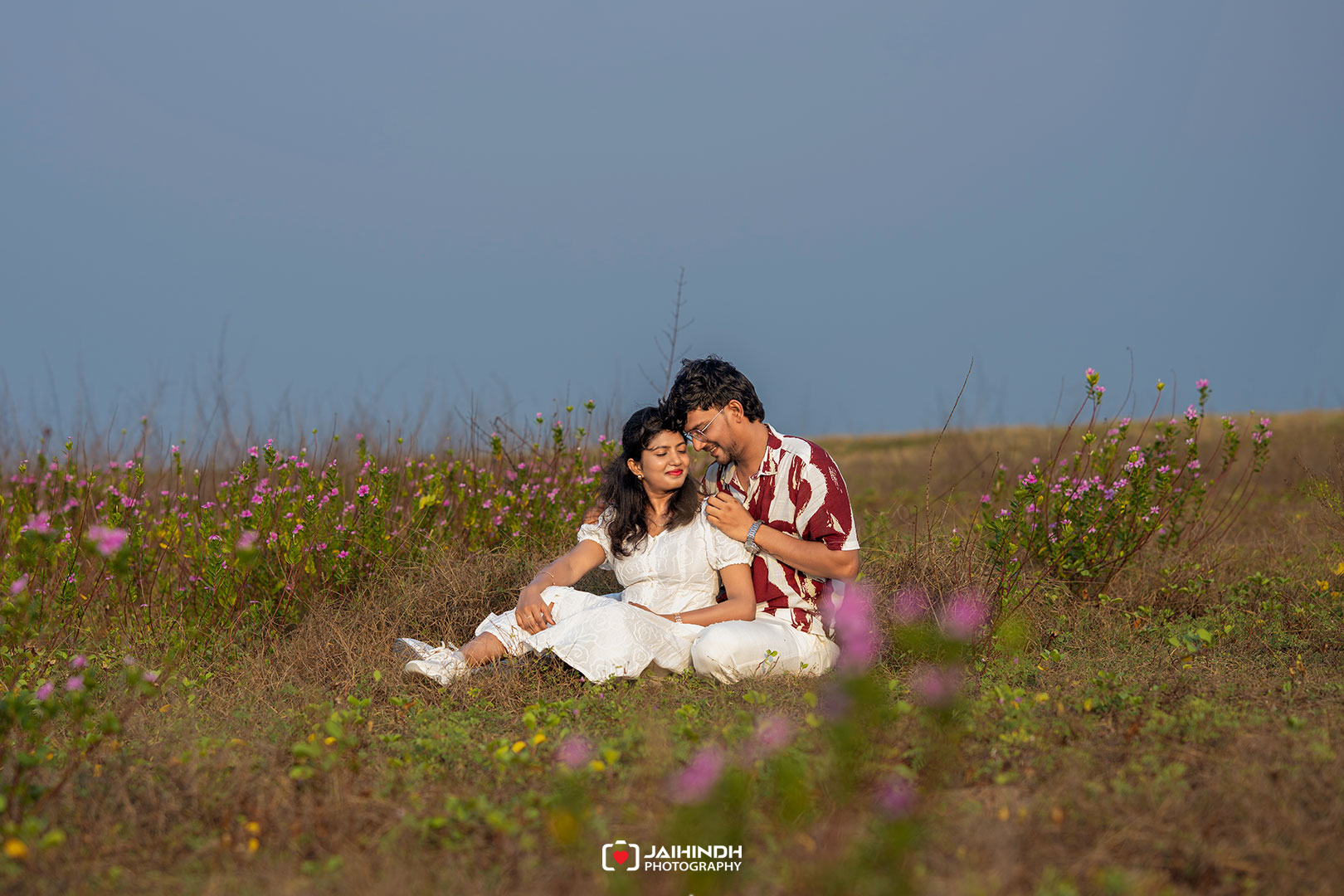 Couple sitting in a wildflower field, leaning against each other and smiling.