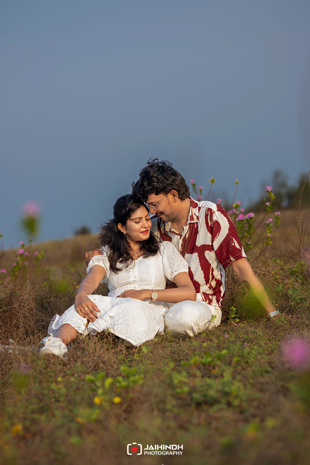 Couple sitting close together in a grassy field with wildflowers, sharing an intimate, peaceful moment outdoors.