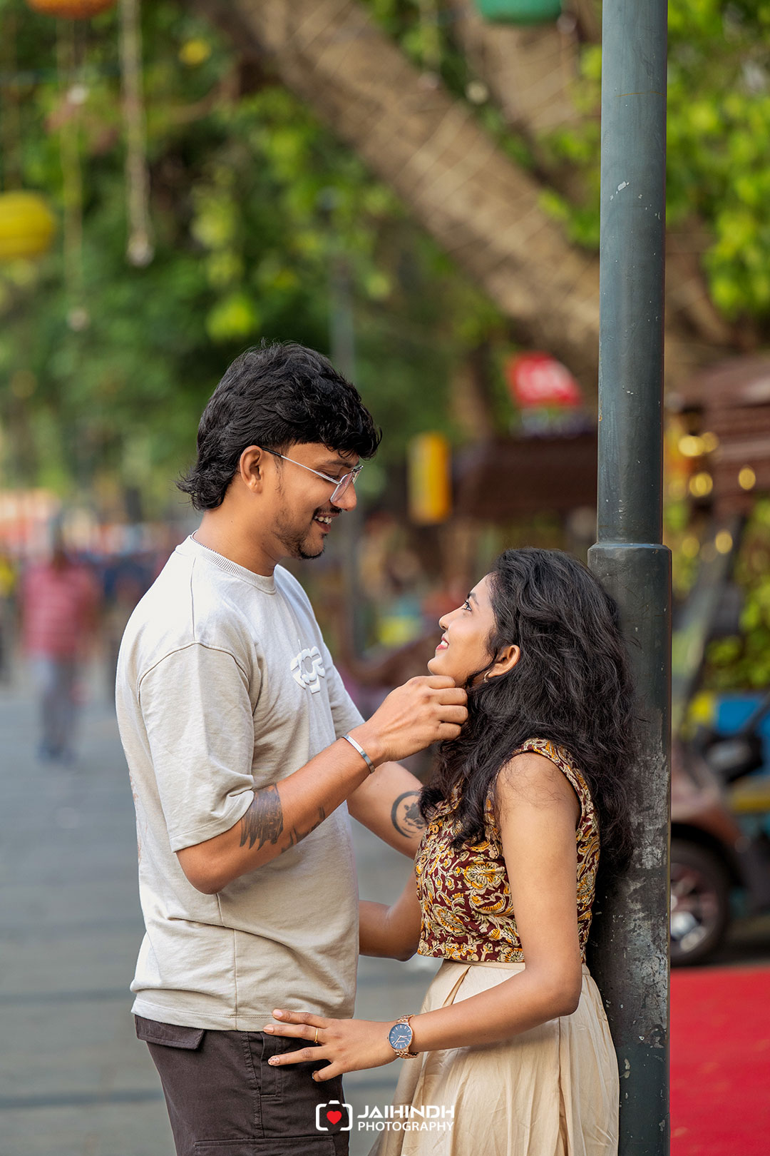Man gently cups the woman's chin as they smile at each other on a city street beside a lamp post, capturing a romantic moment.
