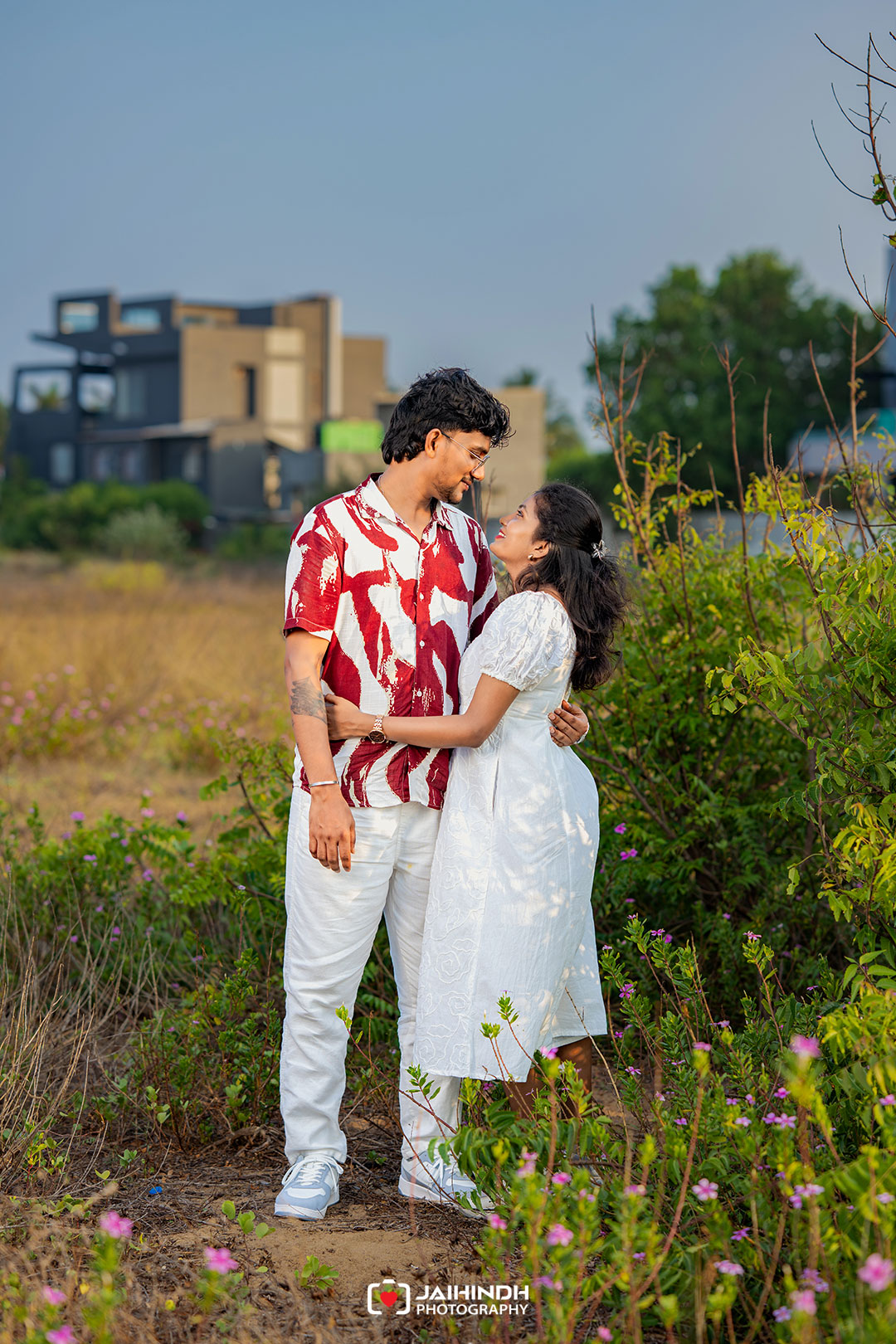 Couple embracing in a grassy field with wildflowers, gazing at each other; modern buildings in the background.
