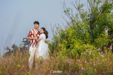 Romantic couple embracing in a field of pink flowers with green shrubs and a clear blue sky behind them.