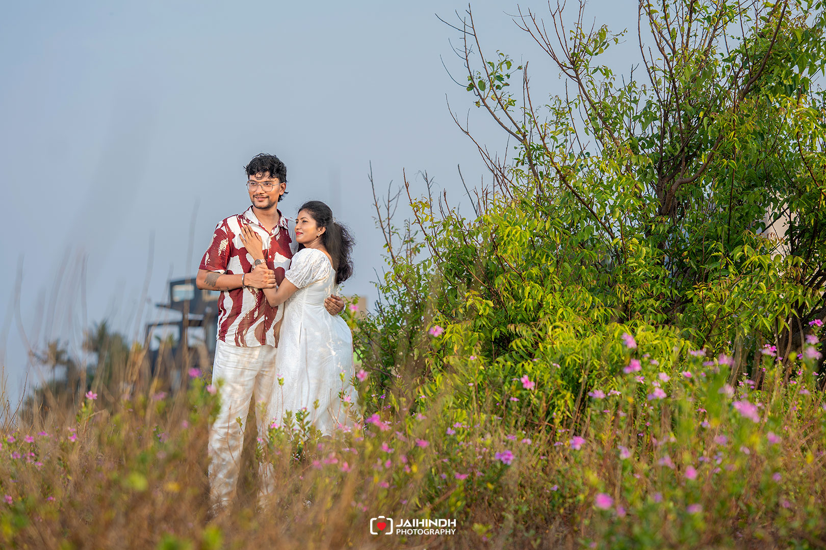 Romantic couple embracing in a field of pink flowers with green shrubs and a clear blue sky behind them.