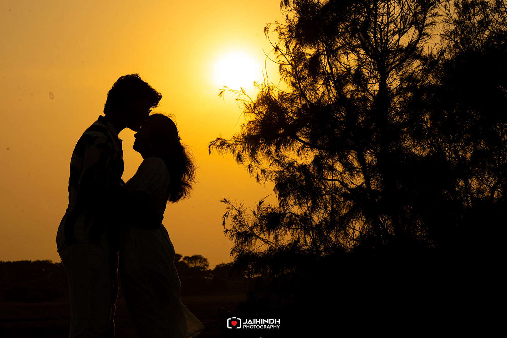 Silhouetted couple kissing at sunset with a tree on the right.