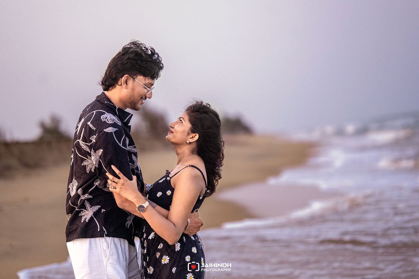Couple embracing on a sandy beach, gazing into each other's eyes as waves roll in at sunset.
