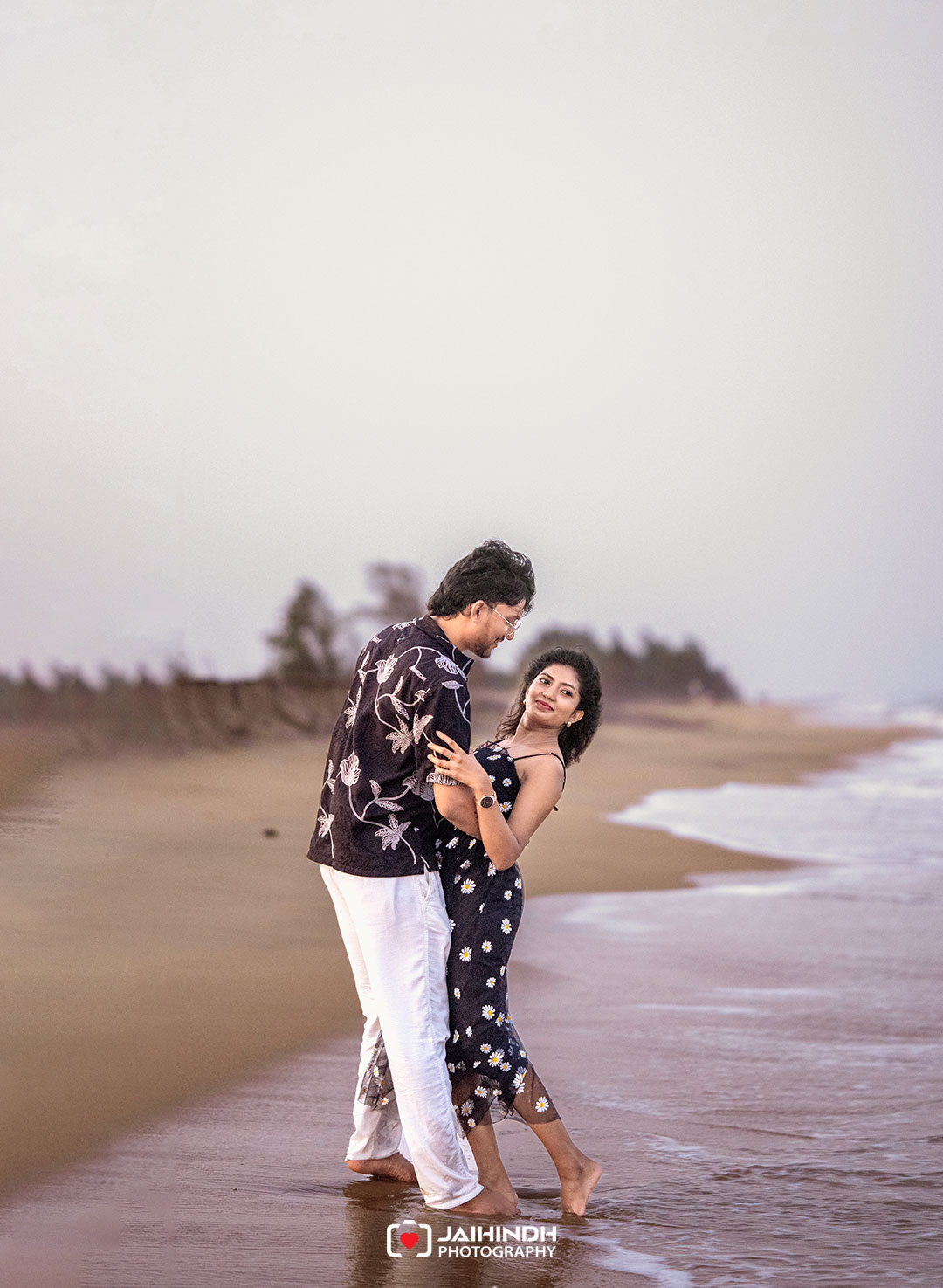 A couple stands barefoot on the wet sand, embracing and smiling at each other along the shoreline.