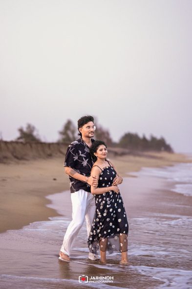 Couple walking along a sandy beach at the shore, the man embracing the woman from behind as waves touch their feet at dusk