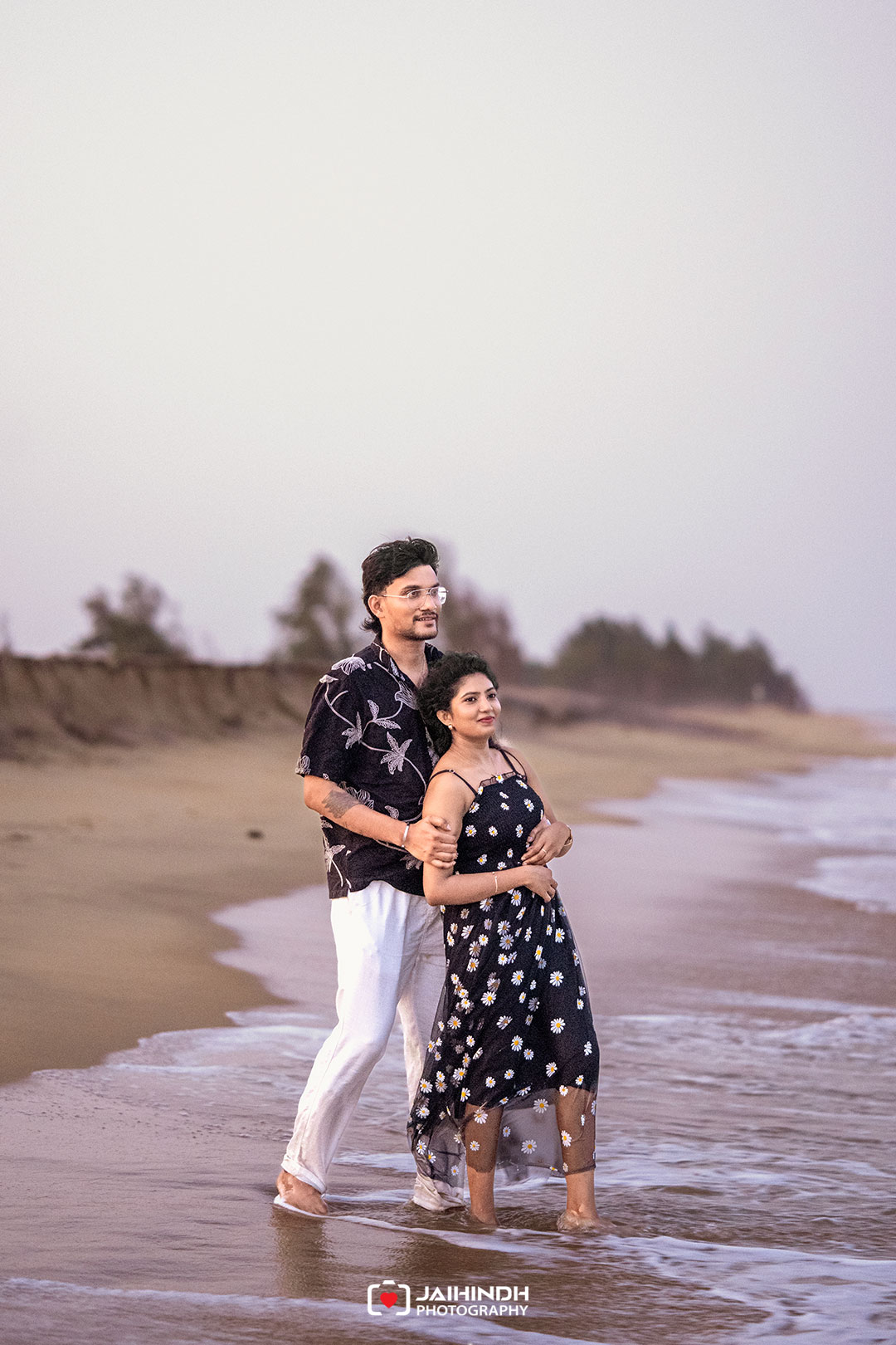 Couple walking along a sandy beach at the shore, the man embracing the woman from behind as waves touch their feet at dusk