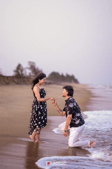 Couple on a beach; man kneels in front of woman, holding her hand as they share a romantic moment by the waves.