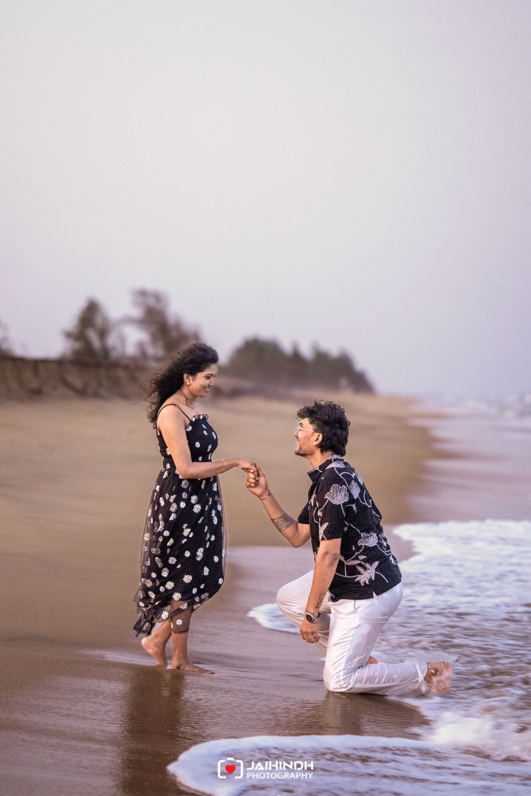 Couple on a beach; man kneels in front of woman, holding her hand as they share a romantic moment by the waves.