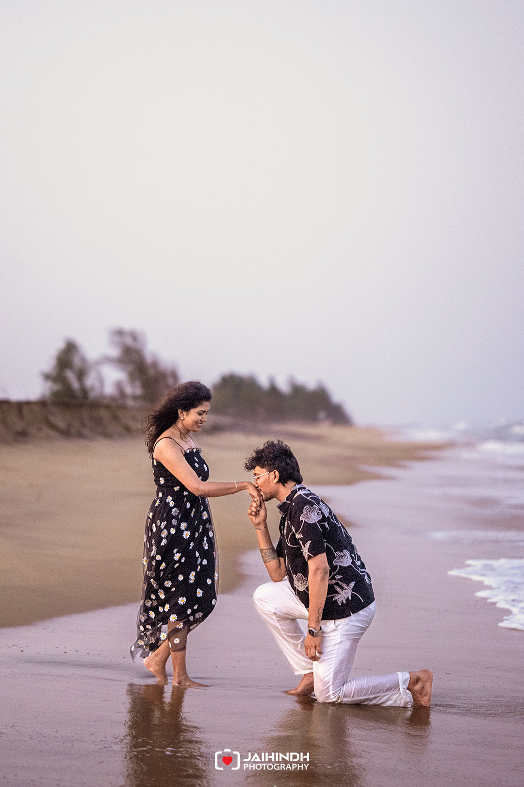 Romantic moment on the beach as a man kneels and kisses a woman's hand by the shore.