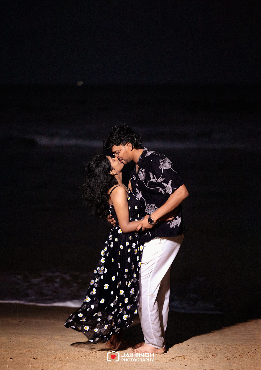 Couple embracing and kissing on a dimly lit beach at night with waves in the background.