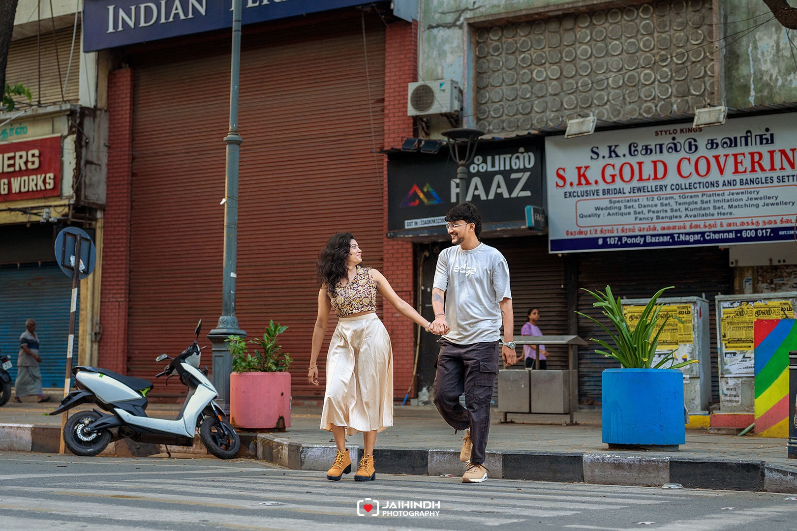 A couple walks hand in hand across a city street, smiling at each other, with shop signs behind them in India.