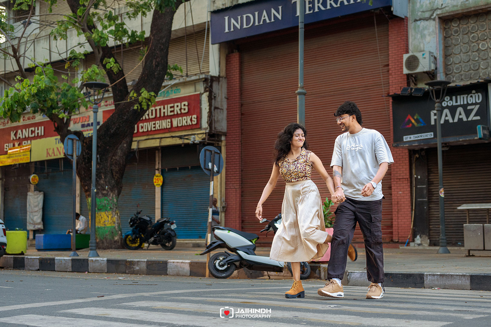 A couple walks hand in hand across a city street as shop shutters are closed behind them, smiling and looking at each other.