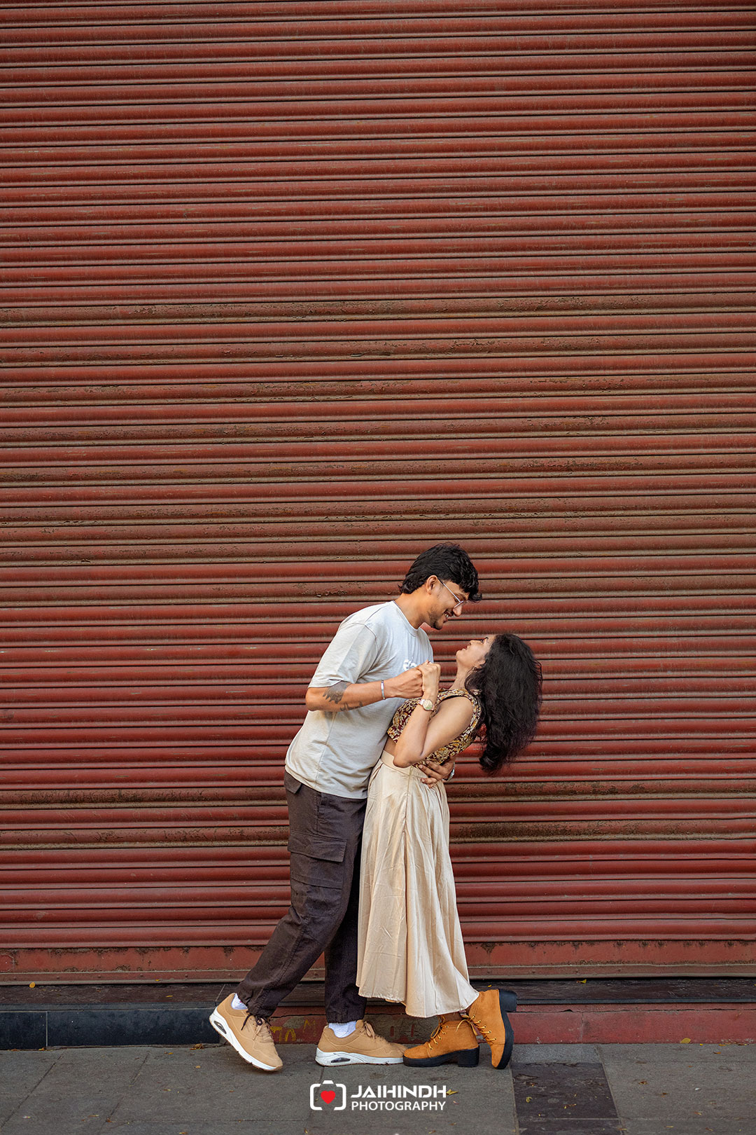 A couple dancing closely in front of a red corrugated metal wall on a city street, holding hands and smiling at each other.