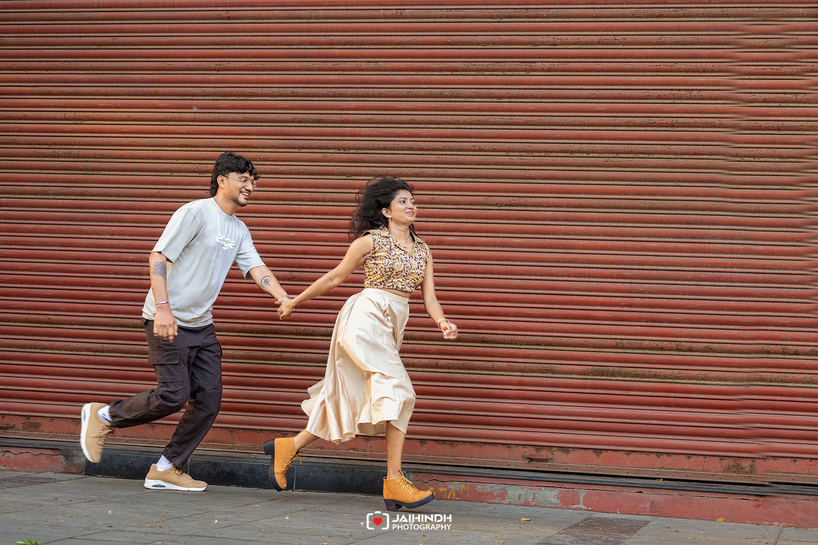 A smiling couple runs hand-in-hand along a sidewalk in front of a red corrugated metal shutter, enjoying a sunny day.