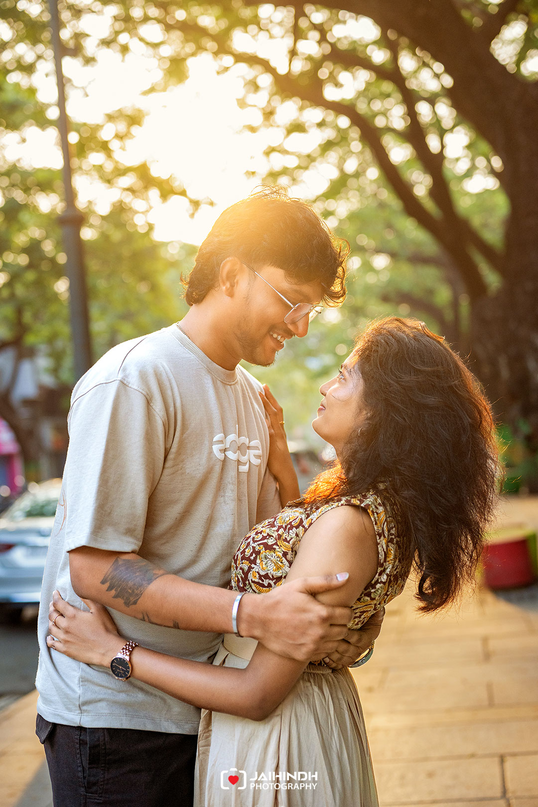 Couple embraces on a sunlit sidewalk, smiling at each other with warm golden hour light.