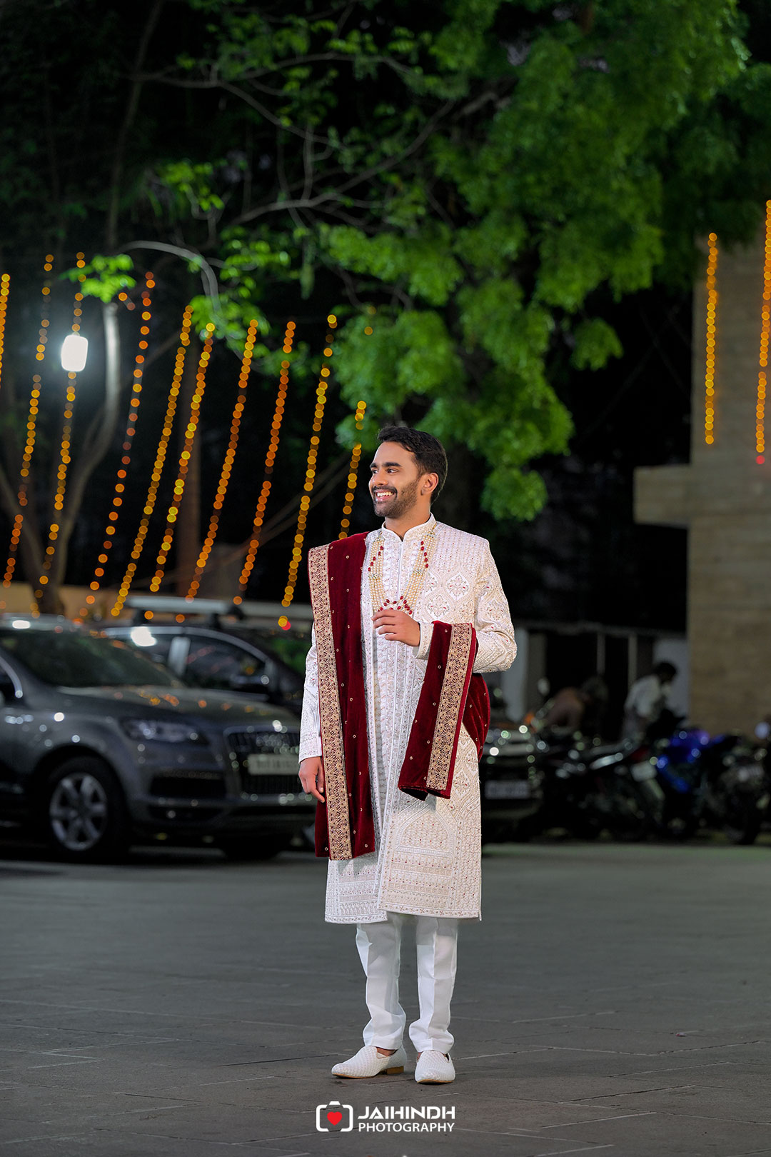 Man in a white traditional sherwani with a maroon embroidered shawl, smiling at night during a festive outdoor event.