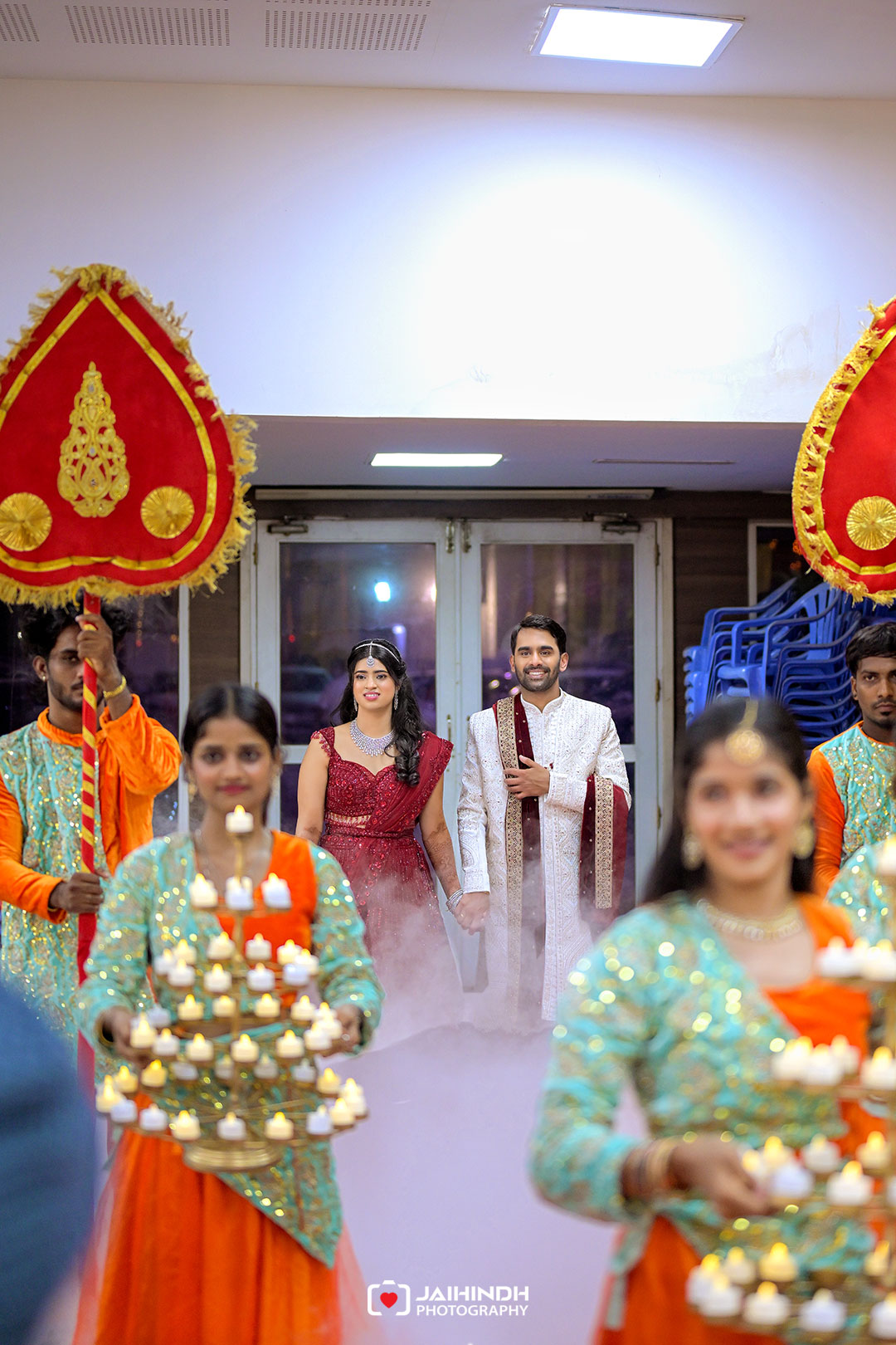 Bride and groom walk hand in hand through a doorway, flanked by dancers carrying lit candles and large red decorative parasols at a festive Indian wedding entrance.