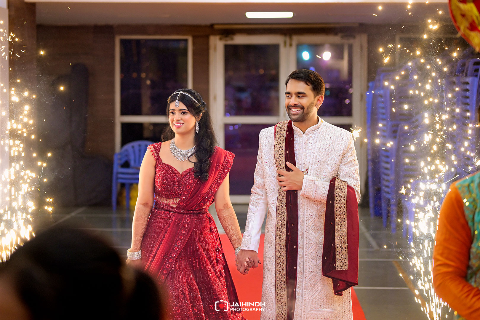 A couple in traditional Indian wedding attire walk hand in hand through a spark-filled doorway, smiling at a celebration.
