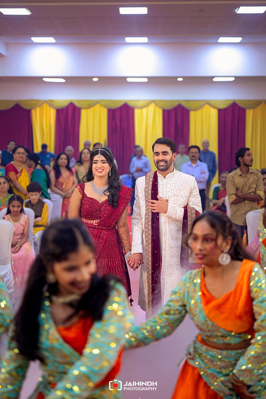 A smiling couple in traditional Indian attire walk hand in hand down an aisle at a wedding reception, surrounded by guests in colorful outfits and yellow-maroon drapes.