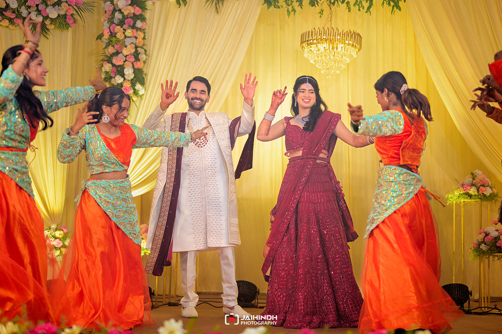 Bride and groom dance on a decorated stage with bridesmaids in turquoise and orange outfits under a chandelier and yellow drapery at a wedding.