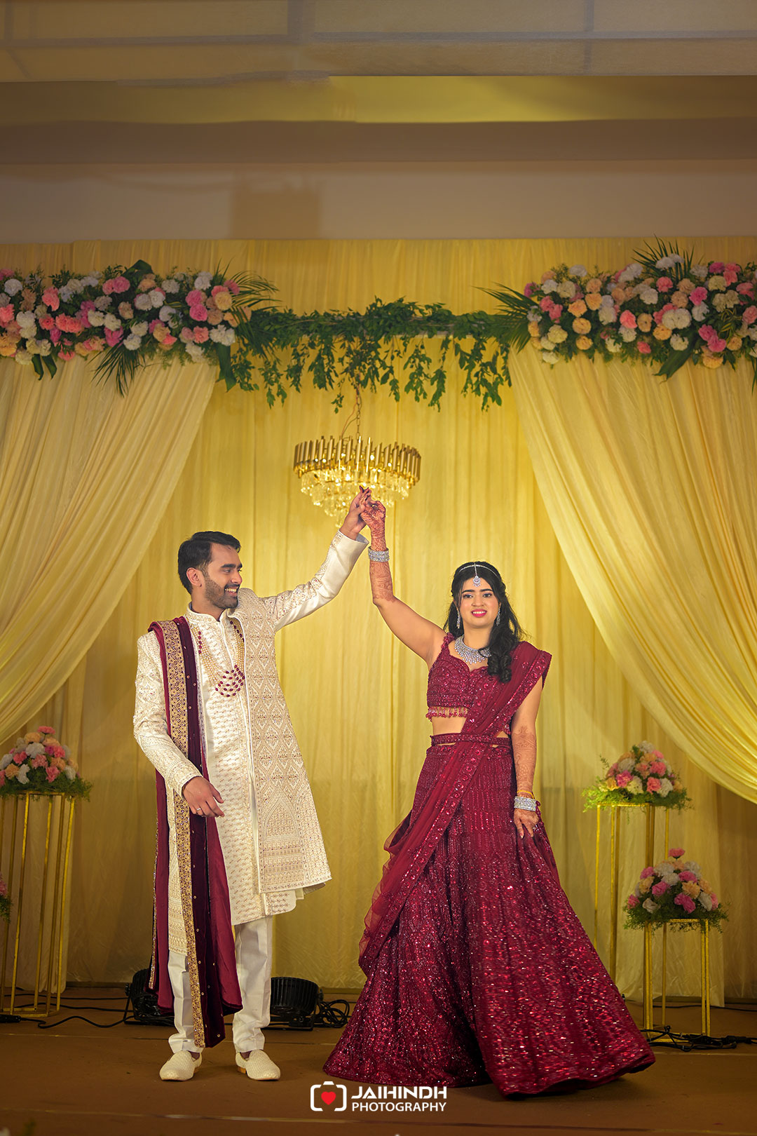 Bride in a maroon lehenga and groom in a cream sherwani raise their hands in a joyful dance on a decorated yellow wedding stage with flowers and chandelier.