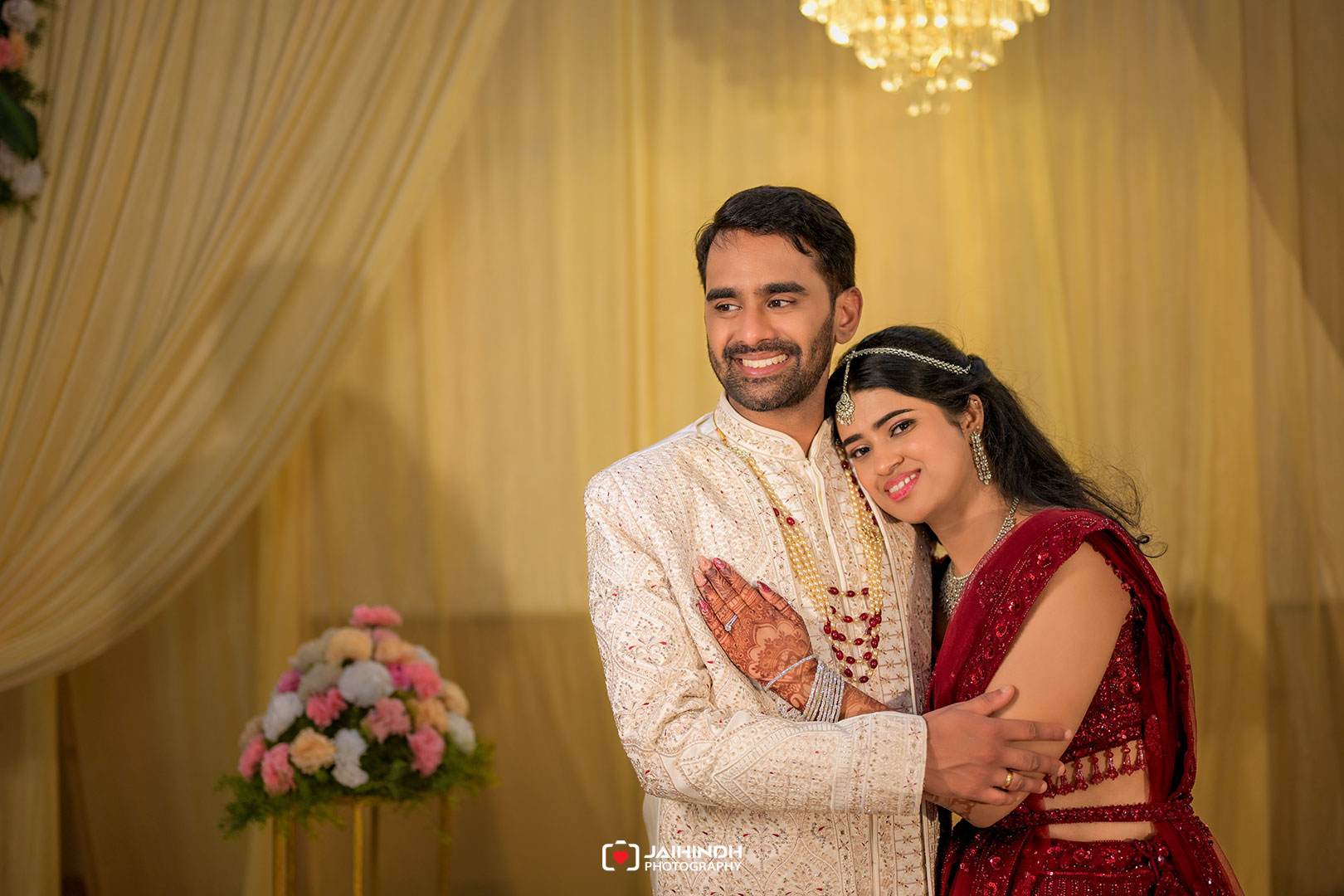 A smiling couple in traditional Indian wedding attire embracing in front of a gold draped backdrop and chandelier.