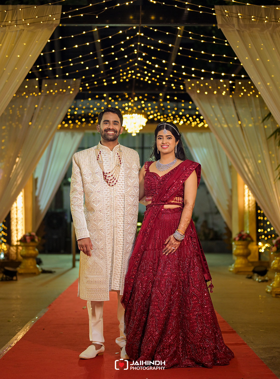 Bride and groom pose together on a red carpet under twinkling string lights and flowing white drapes at a wedding venue; she wears a burgundy lehenga, he a cream sherwani with jewelry.