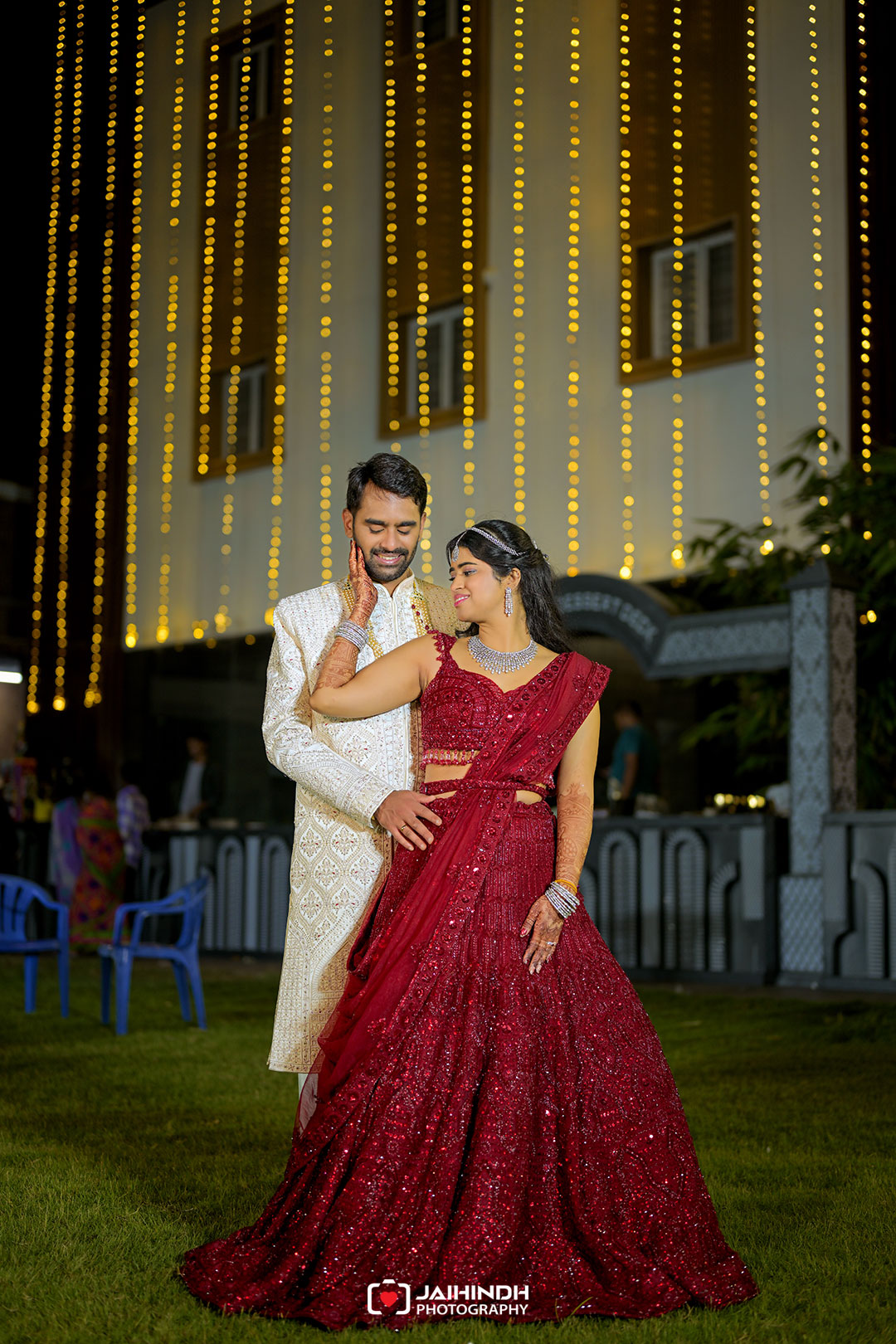 Bride in a red embellished lehenga and groom in a cream sherwani embrace on a lawn at night under vertical string lights.