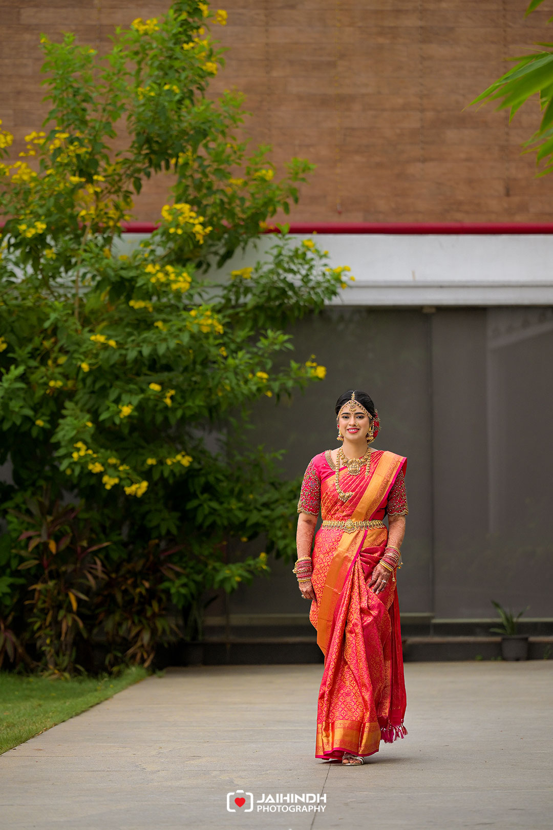 Woman in a pink and orange saree walks along a garden path, smiling, with a flowering bush in the background.