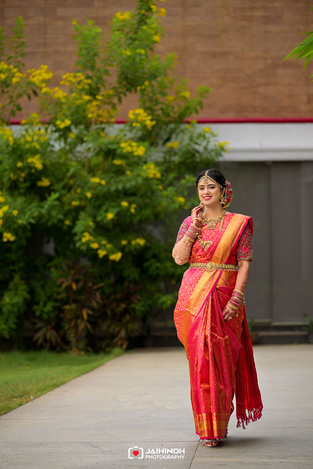 Indian bride in a pink and orange sari with gold jewelry, standing on a walkway outdoors.