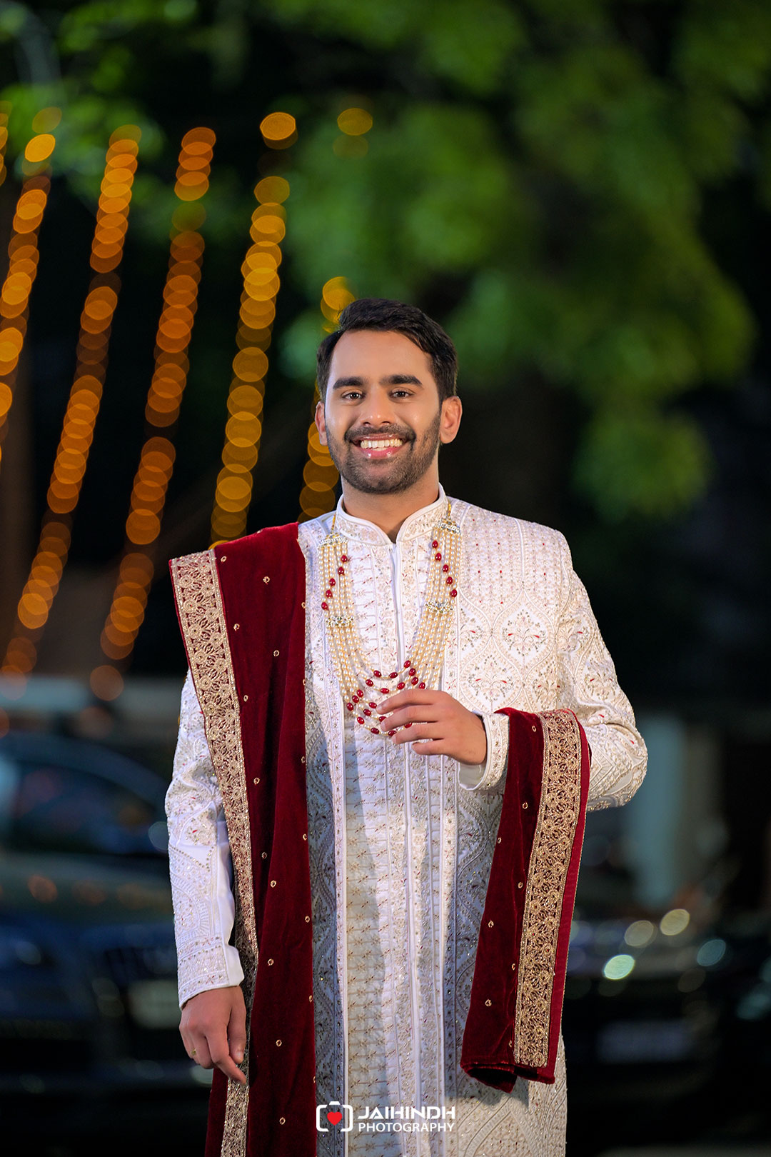 Smiling man in a white ornate sherwani with a red velvet stole and beaded necklace, outdoor wedding backdrop with warm bokeh lights.