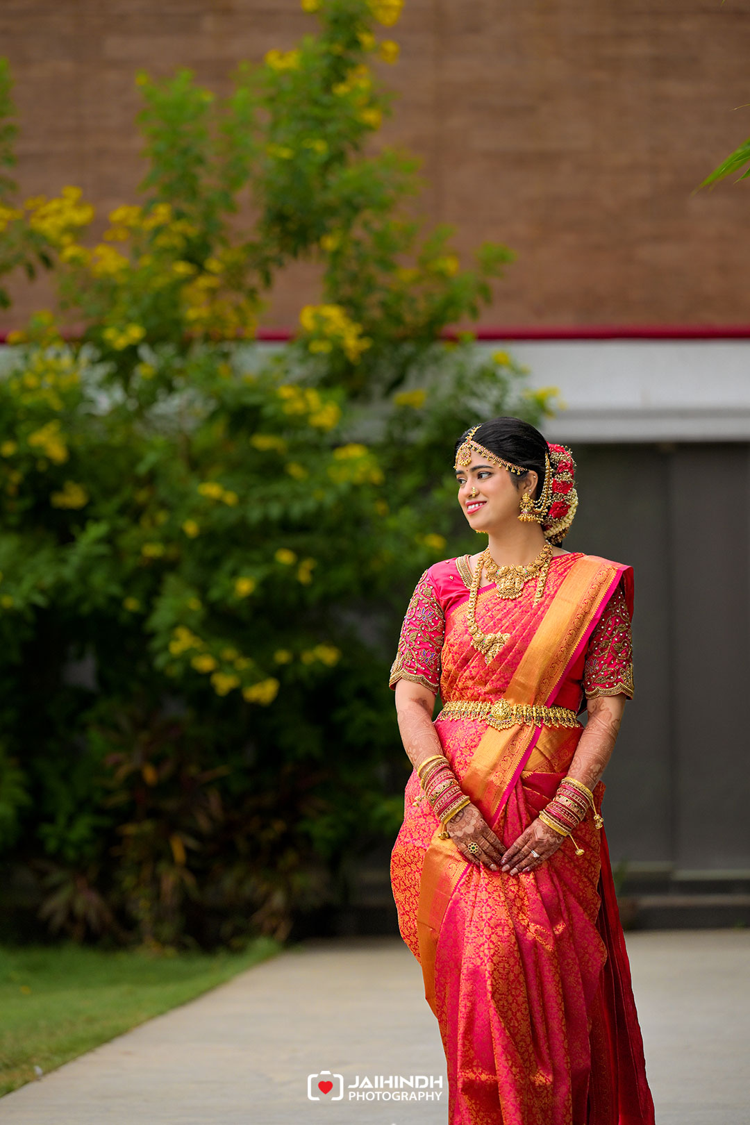 Indian bride in a pink and gold saree, ornate jewelry, and flower-edged bun standing outdoors with greenery in the background.