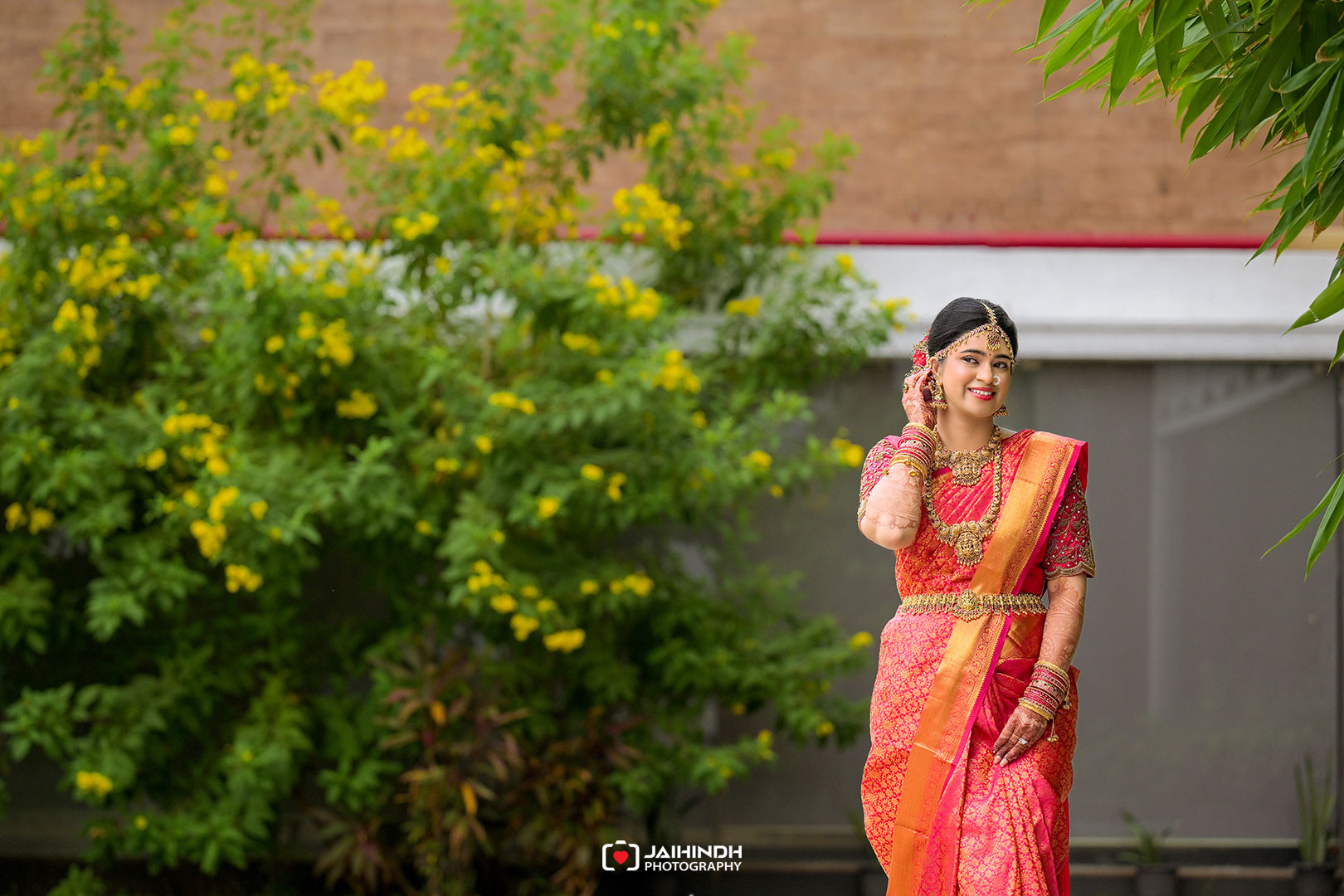 Bride in a pink and orange saree with gold jewelry, posing outdoors among greenery with a smile.