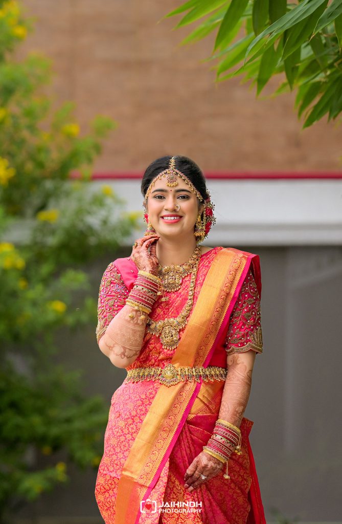 Bride wearing a pink and gold saree with heavy jewelry, smiling outdoors.