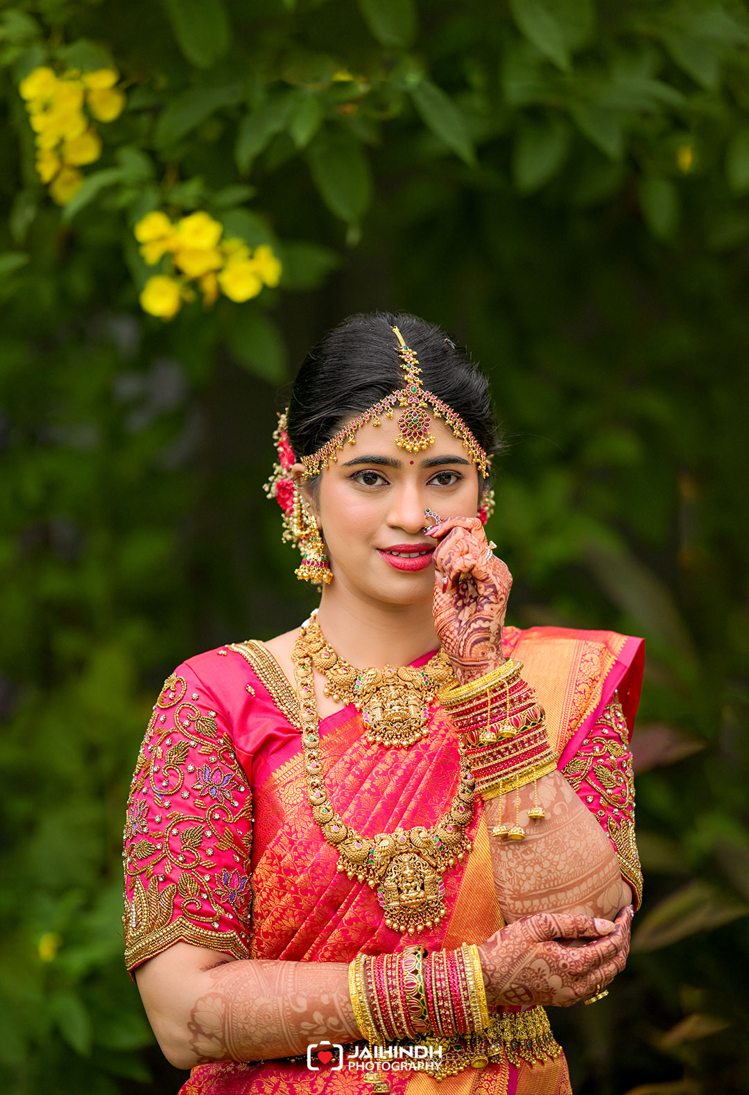 Bride in a pink-orange sari with intricate gold jewelry and mehndi, posing against a lush green garden backdrop.
