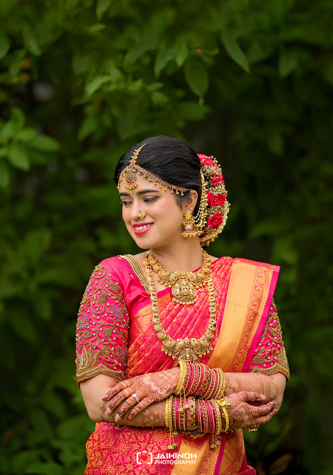 Bride in a pink saree with ornate gold jewelry, hennaed hands, and a floral hair bun, smiling against green foliage.