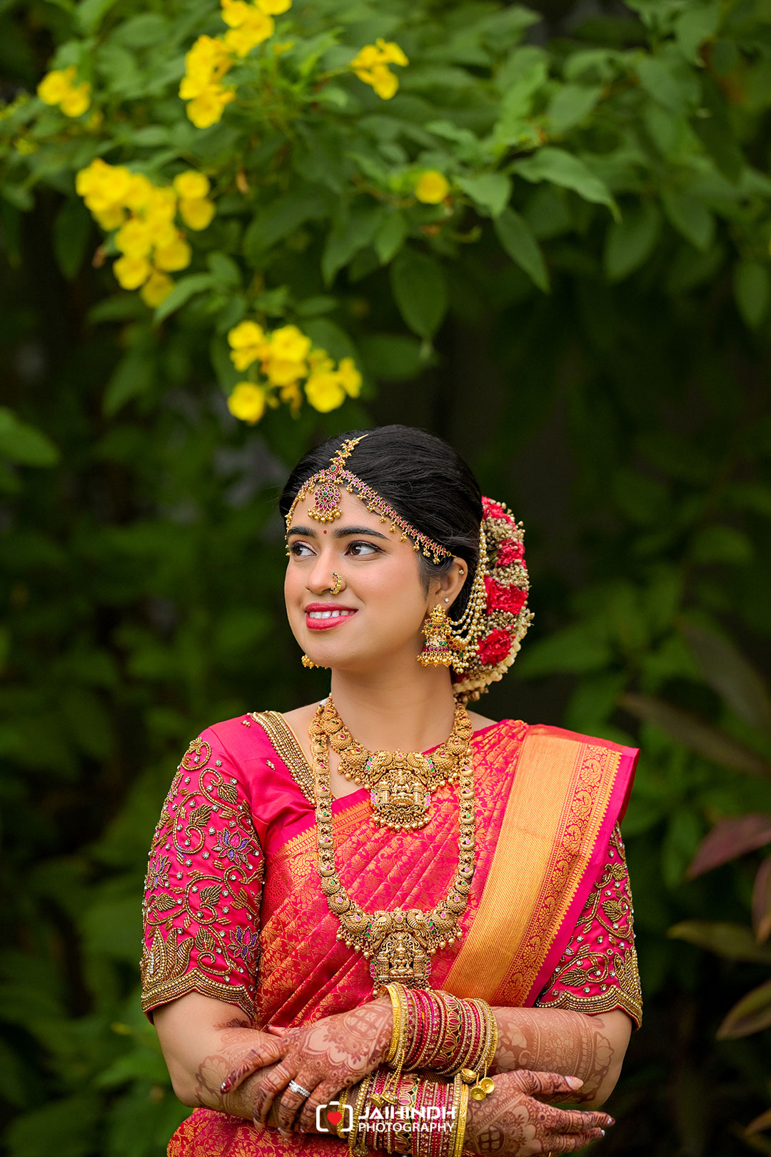 Young Indian bride in a pink saree wearing heavy gold jewelry, with a floral bun, outdoors among green foliage and yellow flowers.