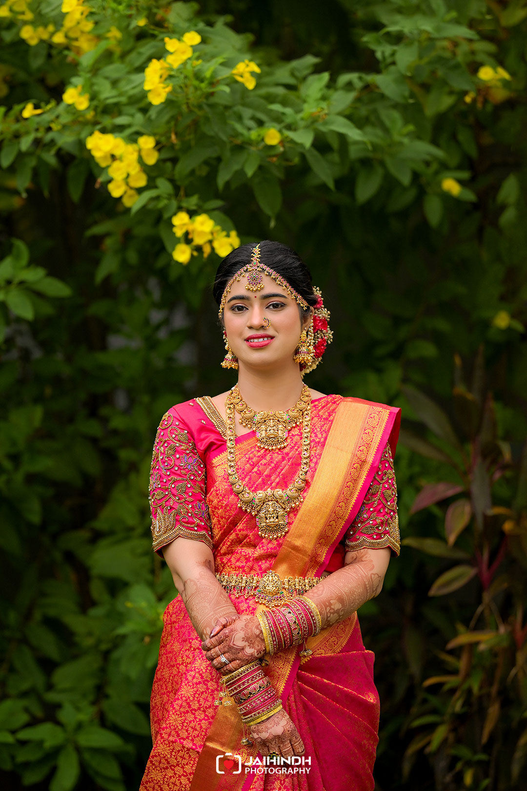 Portrait of a woman in a pink and gold saree wearing heavy traditional jewelry, standing among green plants with yellow flowers in the background, outdoor setting.