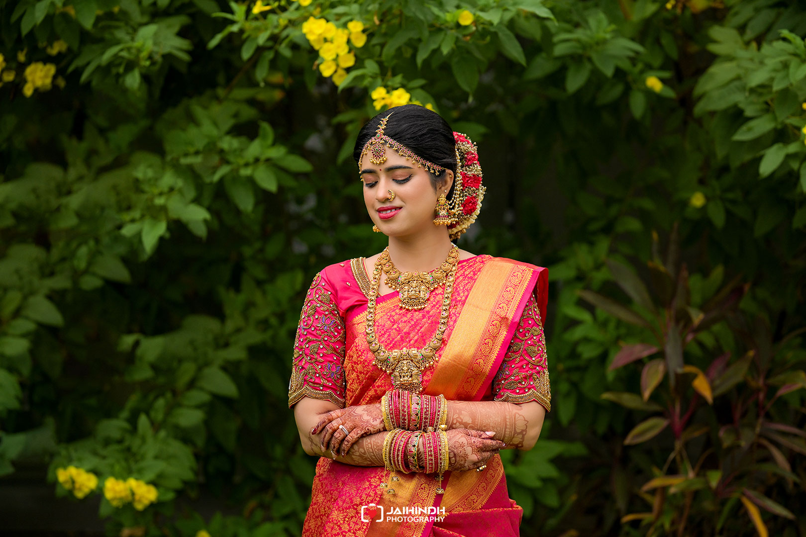 Bride in a pink and gold saree with heavy jewelry and flower bun, posing in a lush garden backdrop.