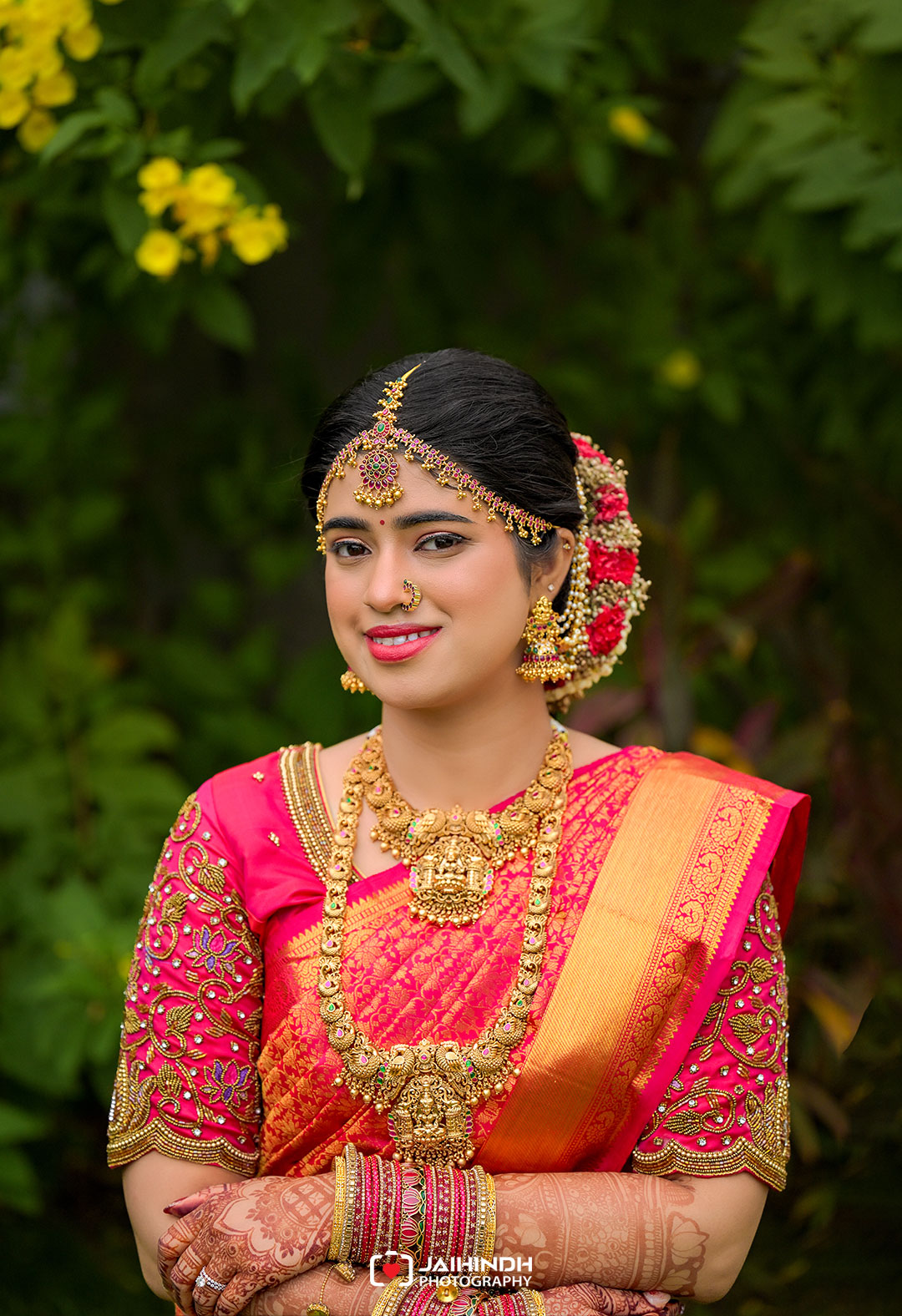 Indian bride in a pink silk saree with gold jewelry, posing outdoors with arms crossed.
