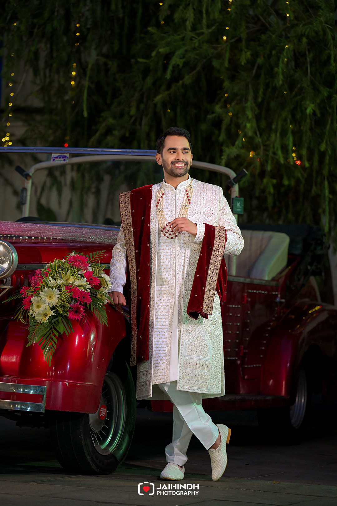 Groom in a white ornate sherwani with a red velvet stole, posing beside a red vintage car with flowers on the hood.