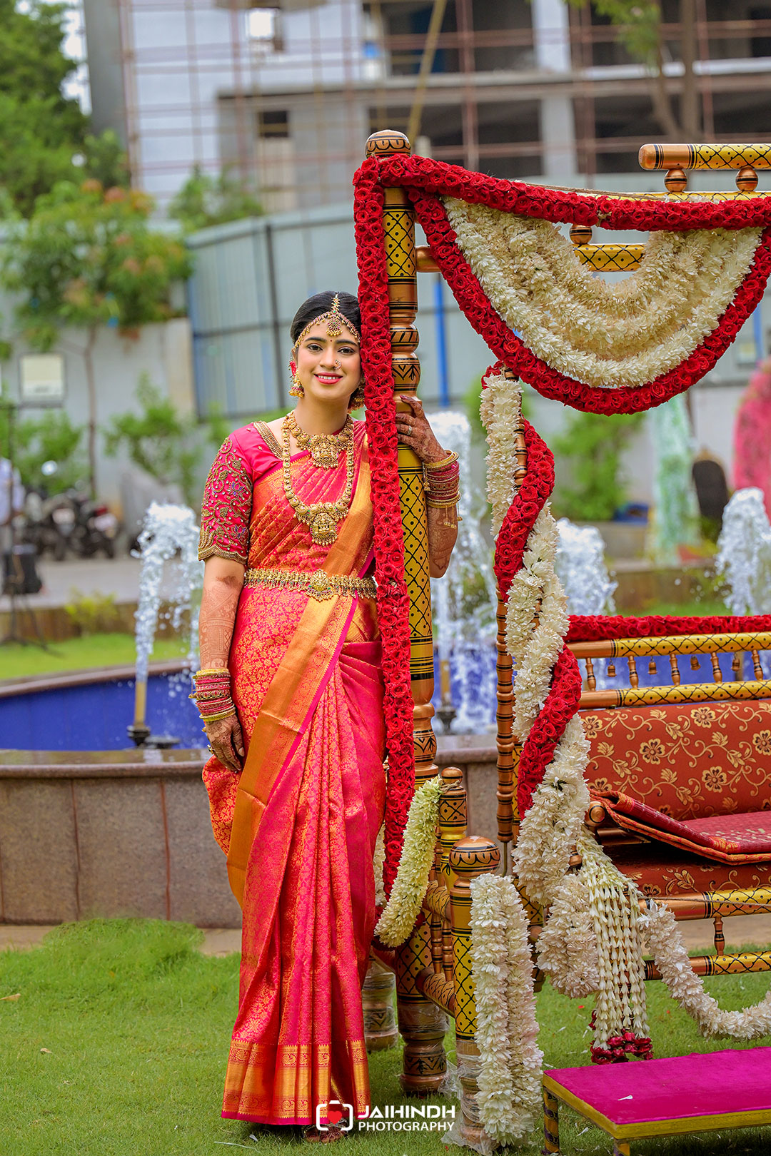 Bride in a pink-orange saree stands beside a flower-decorated swing at an outdoor Indian wedding.