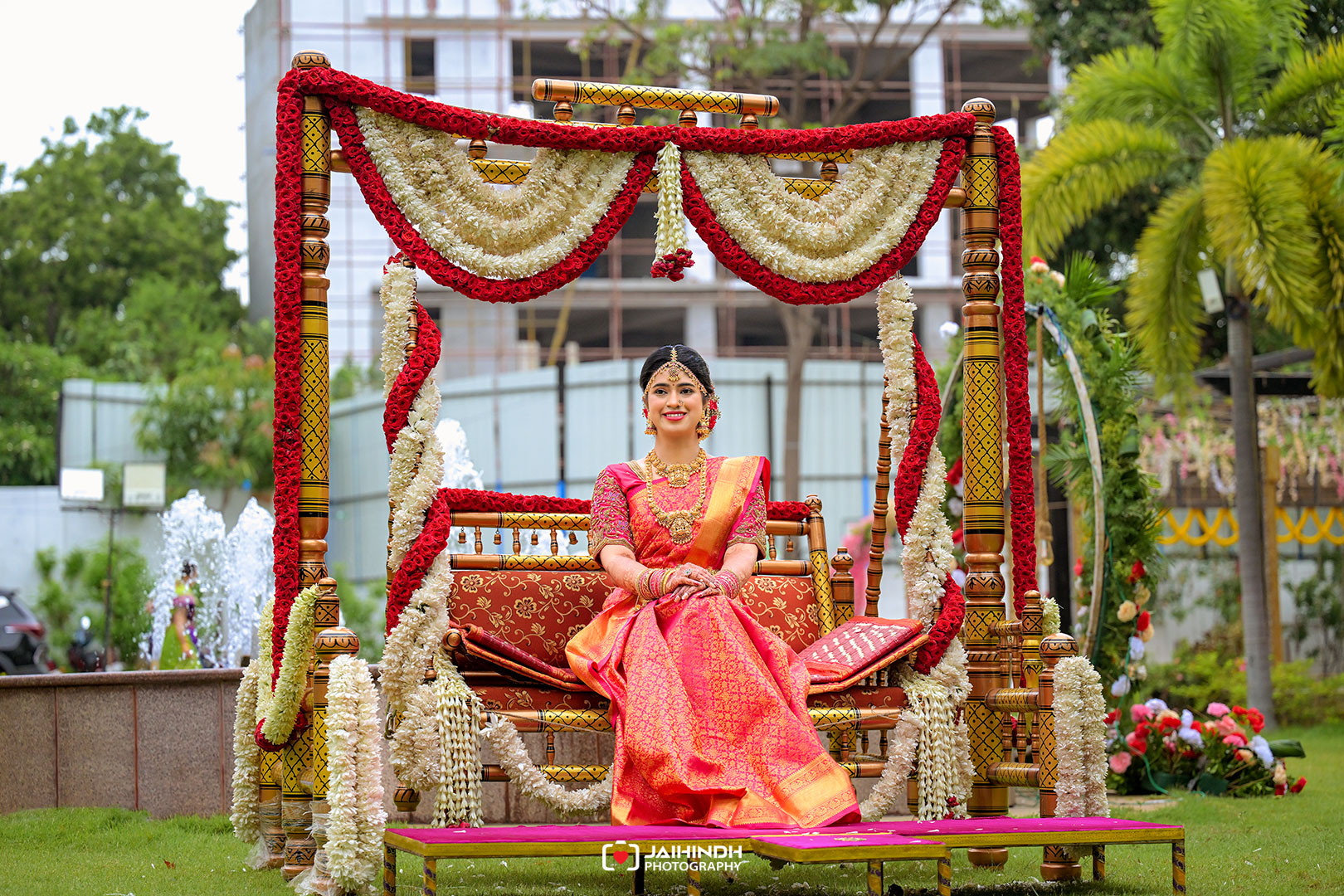 Bride in an orange-pink saree sits on a flower-decorated swing at an outdoor wedding venue with jewelry and red-gold attire.