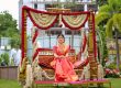 Woman in a pink-orange sari sits on an ornate flower-decorated swing outdoors, surrounded by red and white garlands for a wedding.