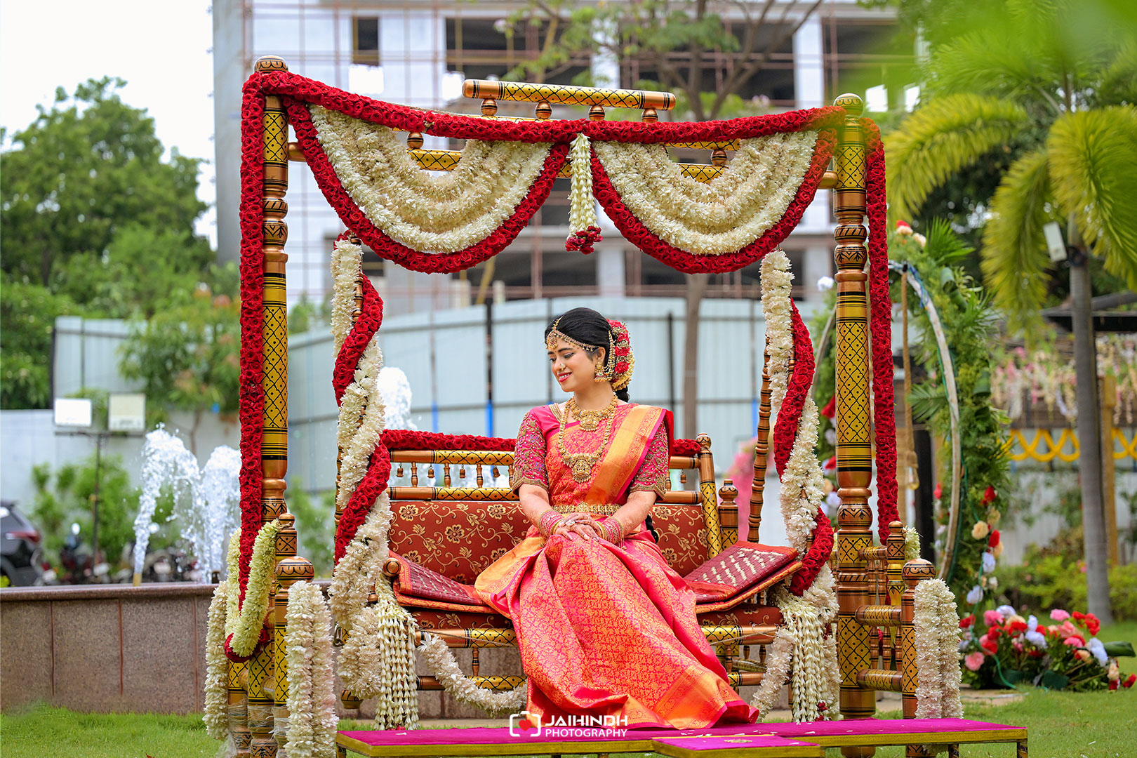 Bride in a pink and gold saree sits on a flower-decorated ceremonial swing outdoors, wearing jewelry and a floral hairpiece.