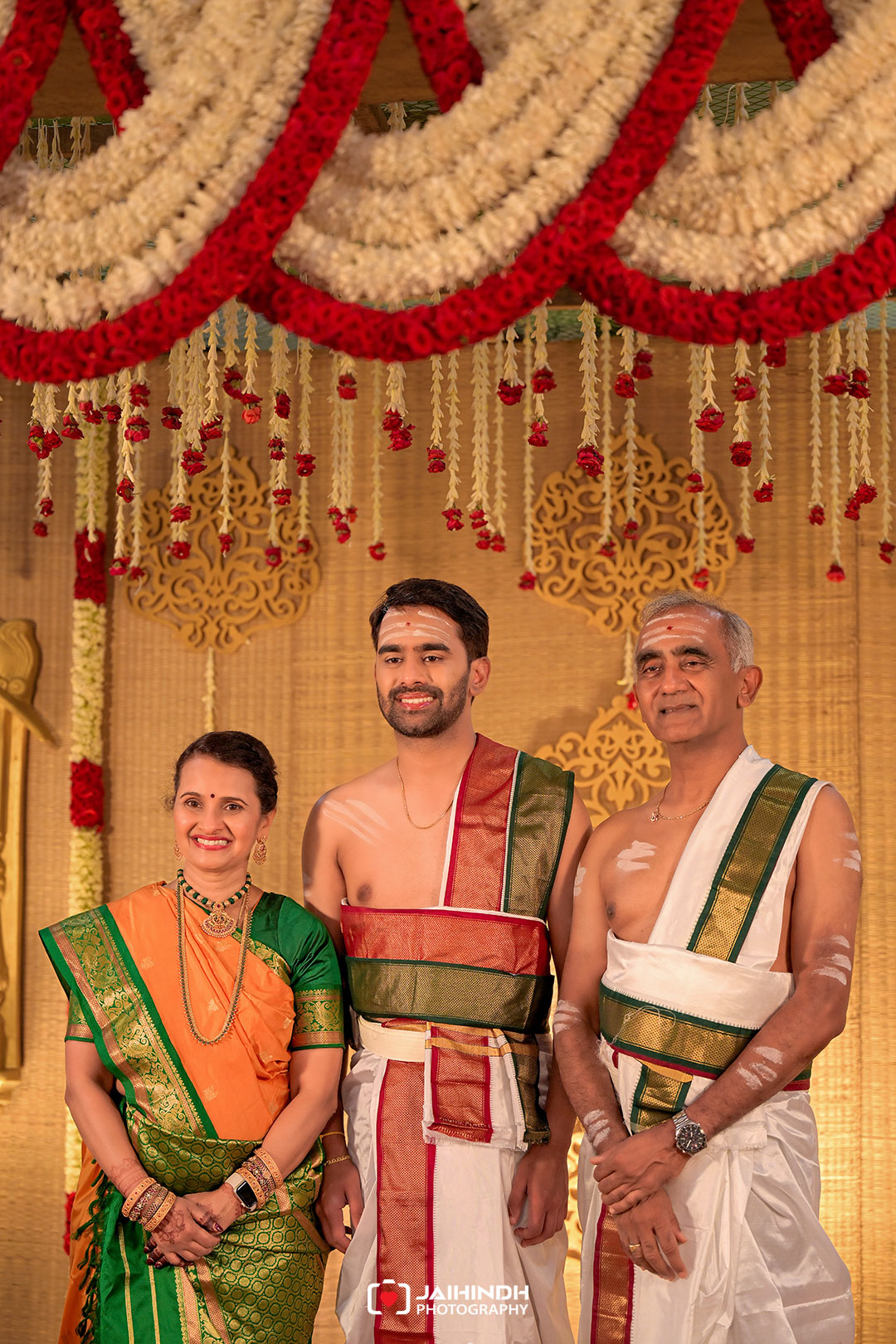 A smiling family of three in traditional Indian attire posing against a decorated backdrop of garlands and ornate patterns.