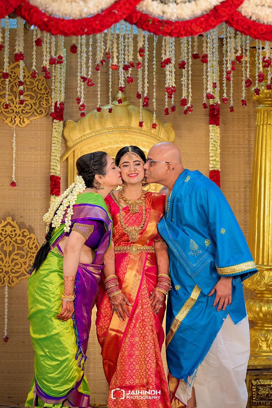 Bride in red and gold saree stands center as two relatives kiss her cheeks on a ornate wedding stage adorned with gold decor and red flowers.
