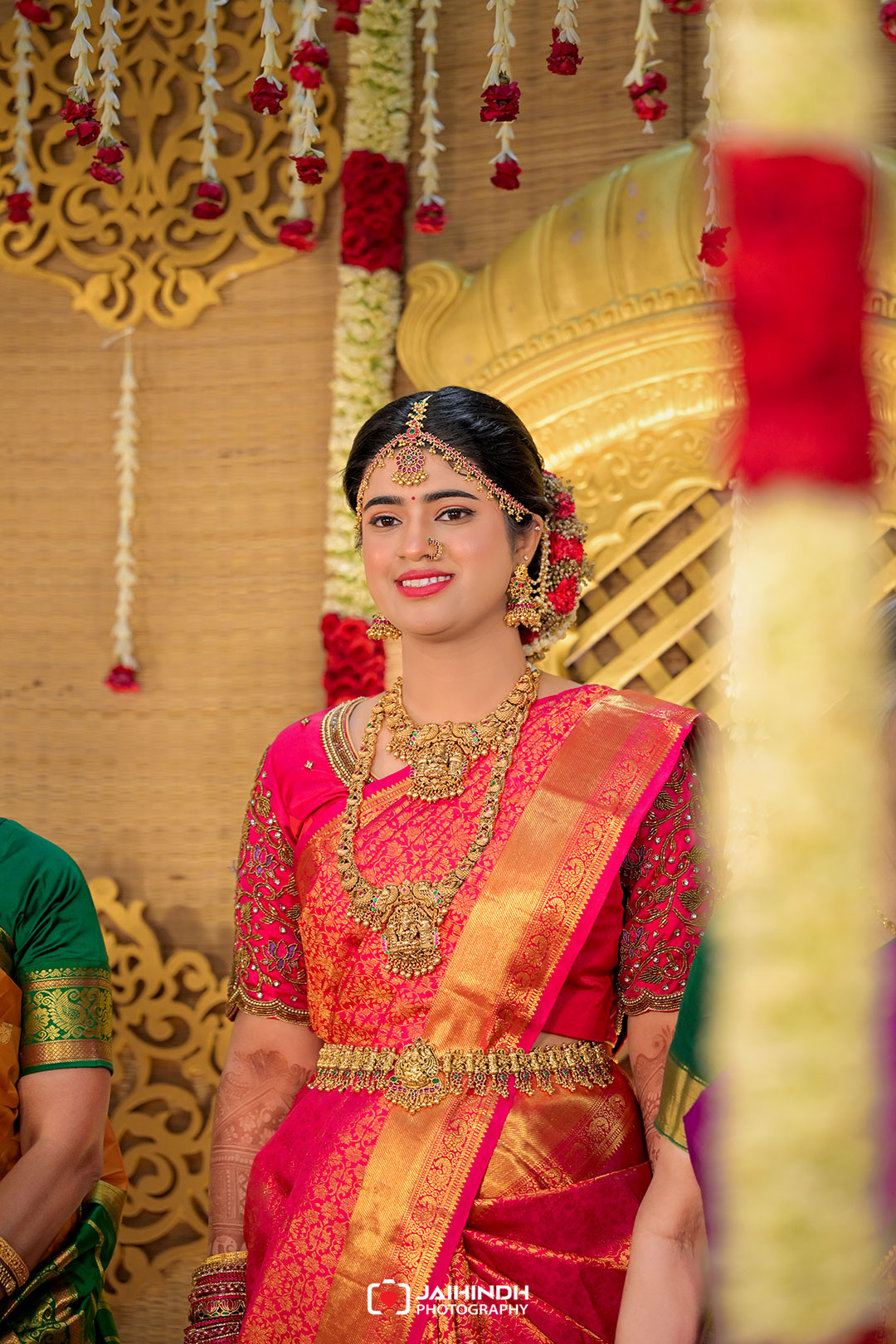 Bride in a magenta saree with heavy gold jewelry, smiling on a traditional wedding stage decorated with marigold garlands and gold motifs.