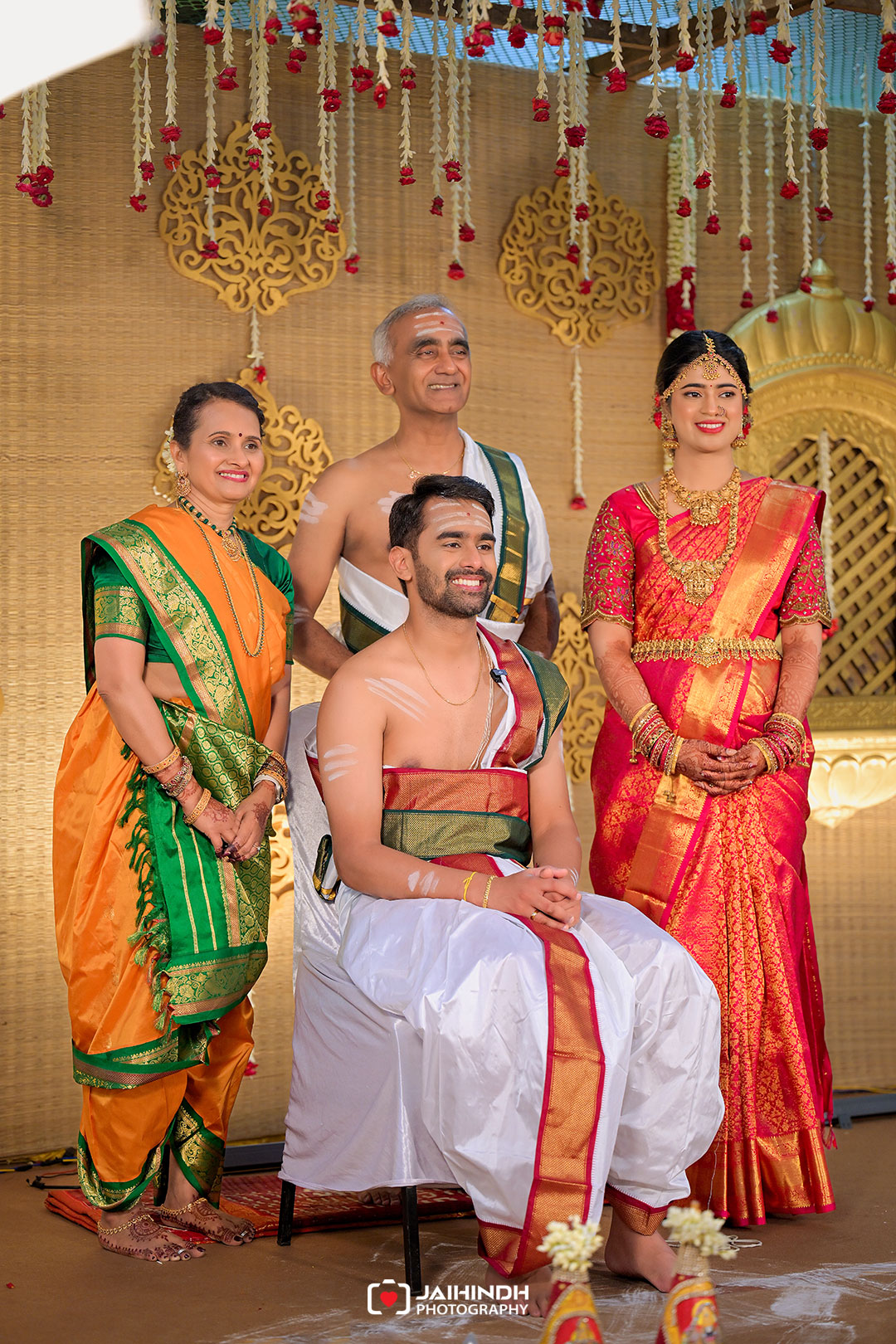 Groom seated in white and gold attire on a stage, flanked by his relatives in colorful traditional saris during a wedding ceremony backdrop with gold decorations.